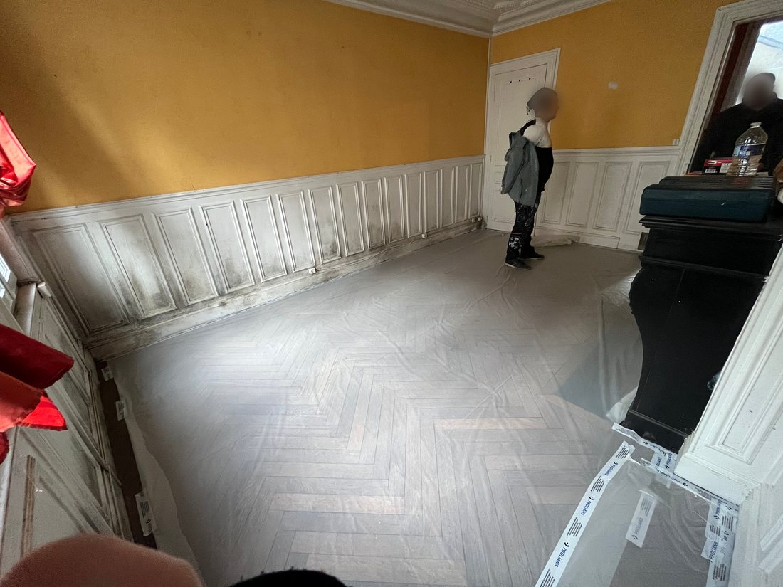 La pièce avant travaux : murs jaunes, boiseries encrassées, parquet point de Hongrie sous bâche, cheminée en marbre noir à droite, deux compagnons sur le chantier.